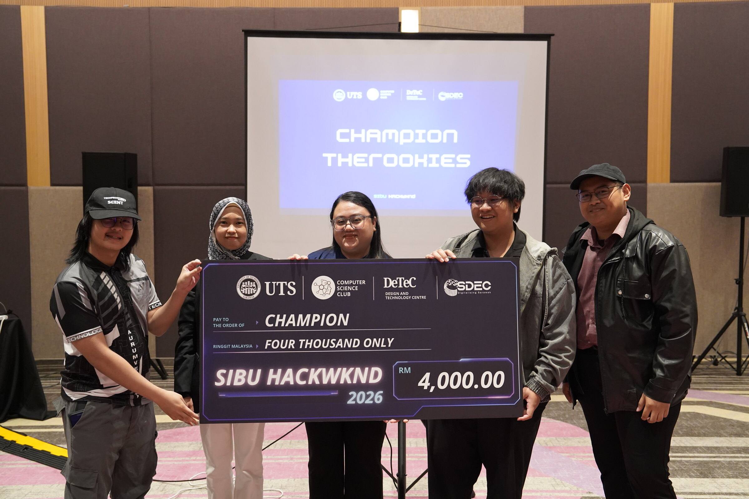 The team in Aurumvelle jerseys holding the Sibu HackWknd 2026 Champion cheque, with Champion TheRookies on the screen behind